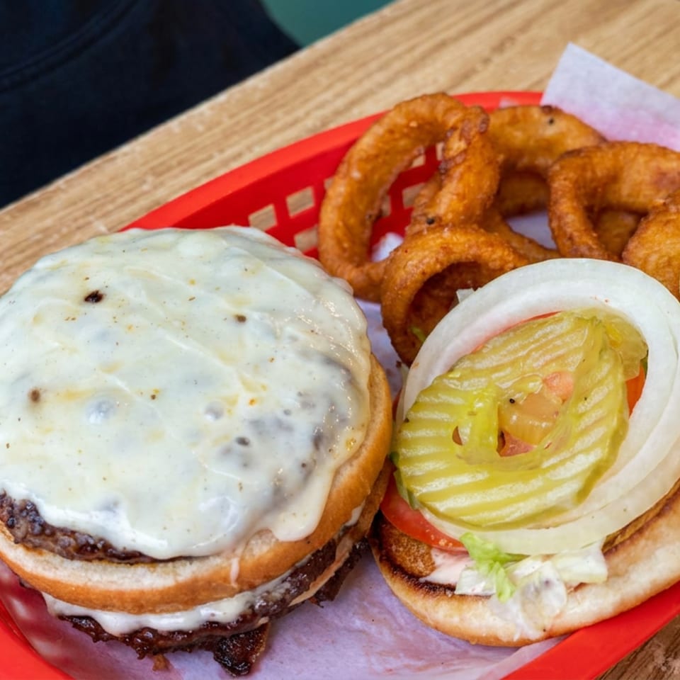 Cheeseburger with lettuce, tomato, onion, and pickle beside fried onion rings on a red basket atop a wooden table.