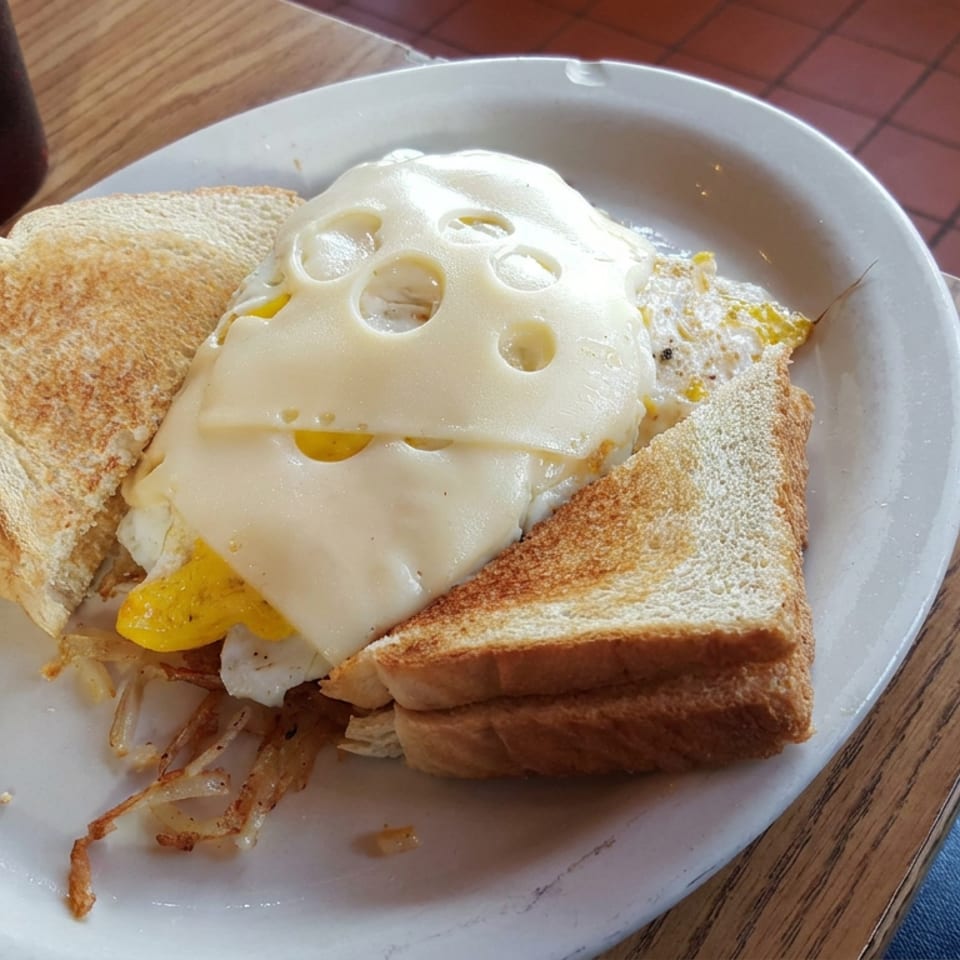 Grilled cheese sandwich layered with fried eggs and melted Swiss cheese on a white plate beside a dark beverage.