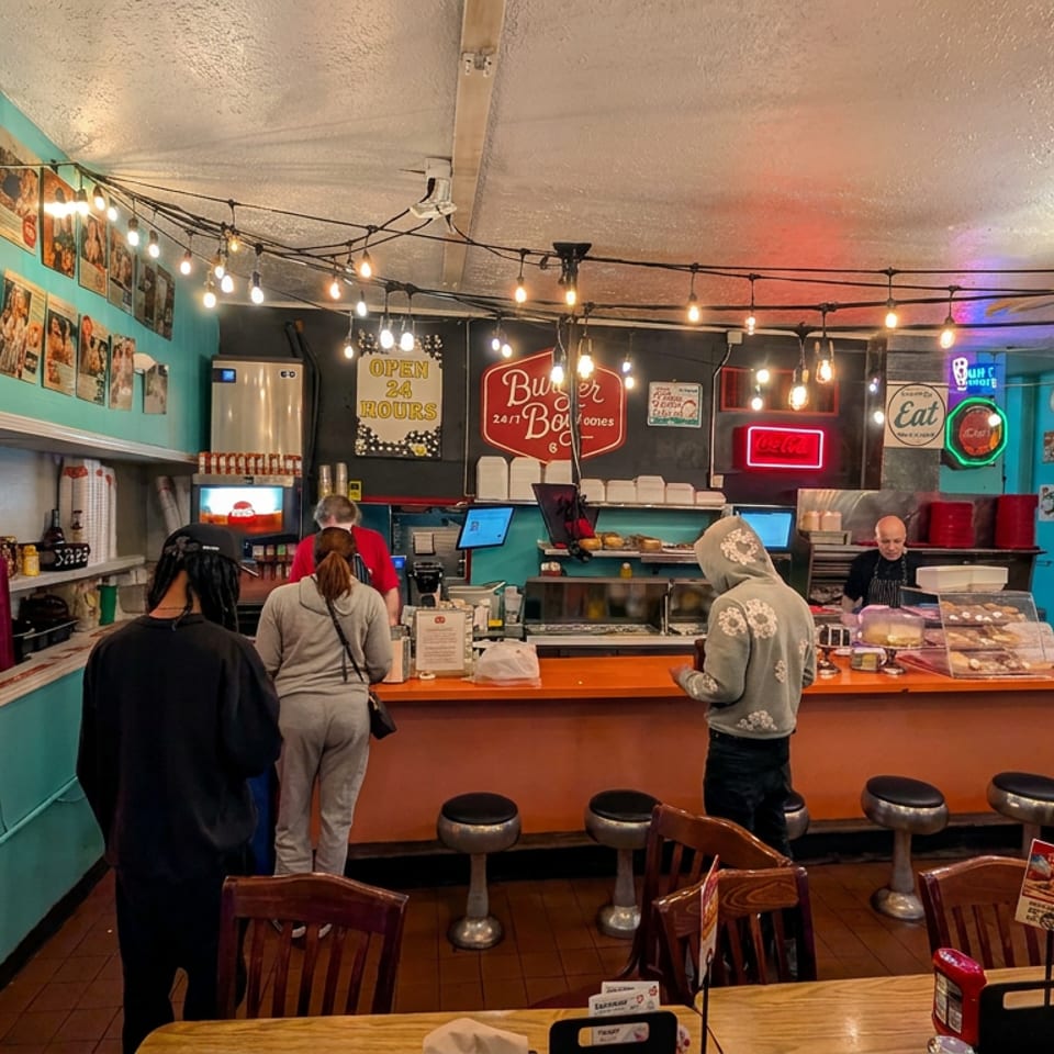 Colorful retro restaurant interior with string lights, counter seating, arcade machine, vintage signs, and customers ordering food.