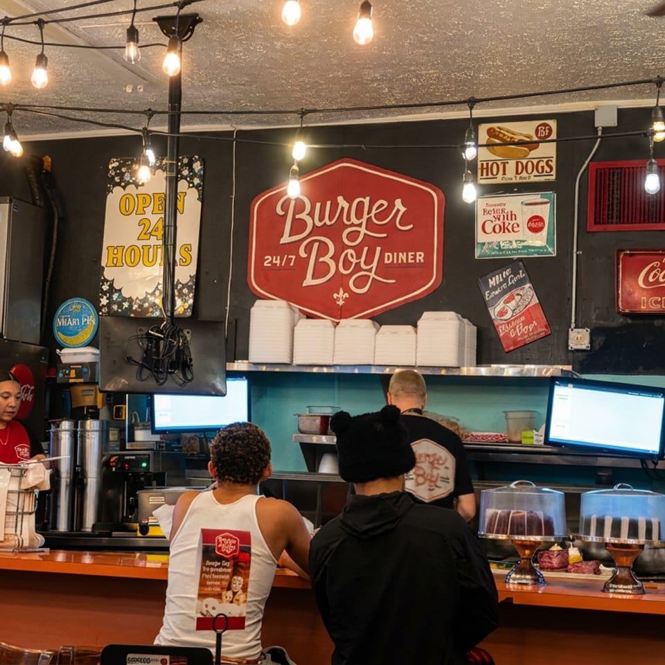 Retro Burger Boy diner counter with staff serving customers, illuminated string lights, vintage signs, and pastries displayed under glass.