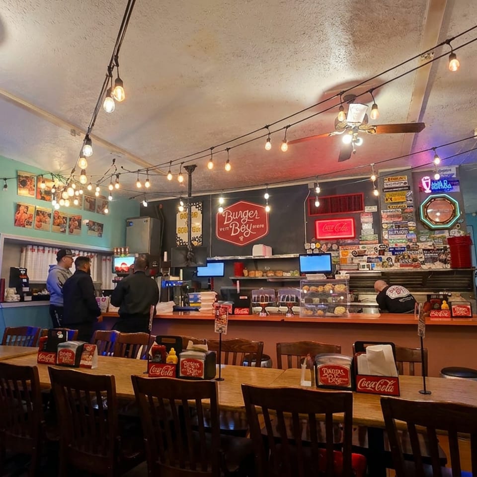 Colorful burger restaurant interior with string lights, wooden tables, counter, retro signs, and several people.