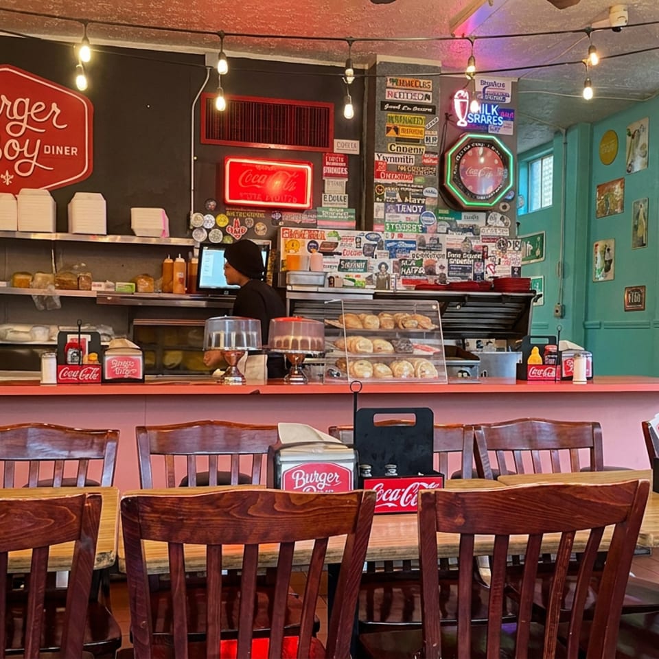 Retro Burger Boy diner interior with wooden tables, counter seating, pastries, soda dispensers, vintage signs, and warm string lights.